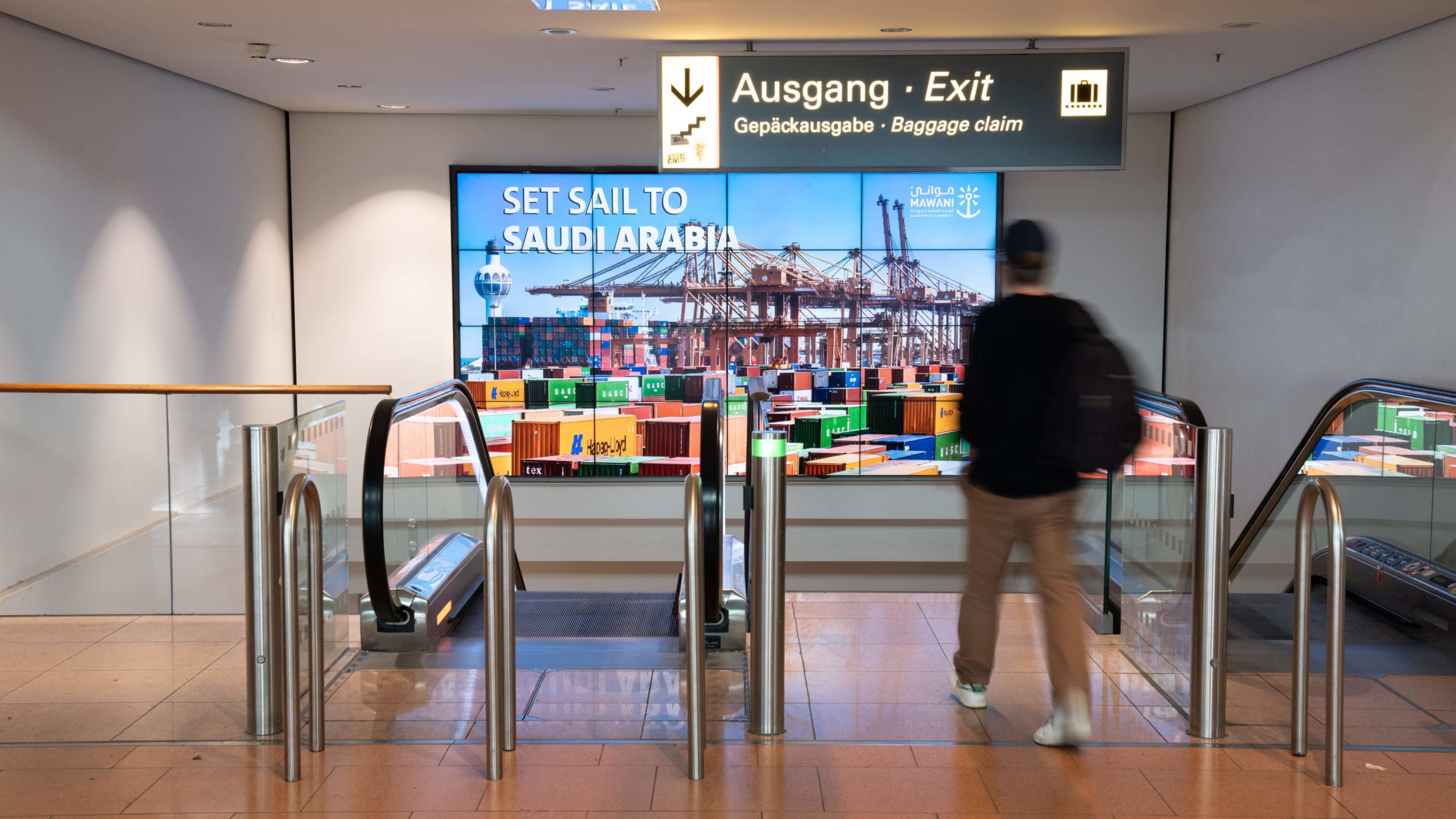 Am Flughafen Hamburg hängt eine digitale Video-Wall an der Wand vor der direkt die Treppen und Rolltreppen Richtung Ausgang hinunterführen. Ein Mann läuft direkt auf die Flughafenwerbung zu, um die Treppe zu nehmen.  