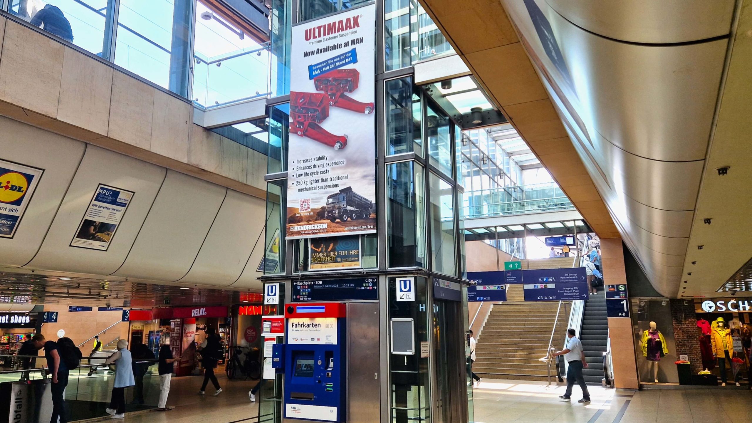 Ein Big Banner der an einem Glasfahrstuhl am Hauptbahnhof in Hannover hängt, wirbt mit der Kampagne von Henrickson. Die Plakatwerbung zeigt ihr neues Produkt. An der Außenwerbung laufen viele Menschen entlang. In Hanover a big banner hanging on a glass elevator at the main station is advertising Henrickson's campaign. The billboard shows their new product. Many people walk past the out-of-home advertising. In Hanover a big banner hanging on a glass elevator at the main station is advertising Henrickson's campaign. The billboard shows their new product. Many people walk past the out-of-home advertising.