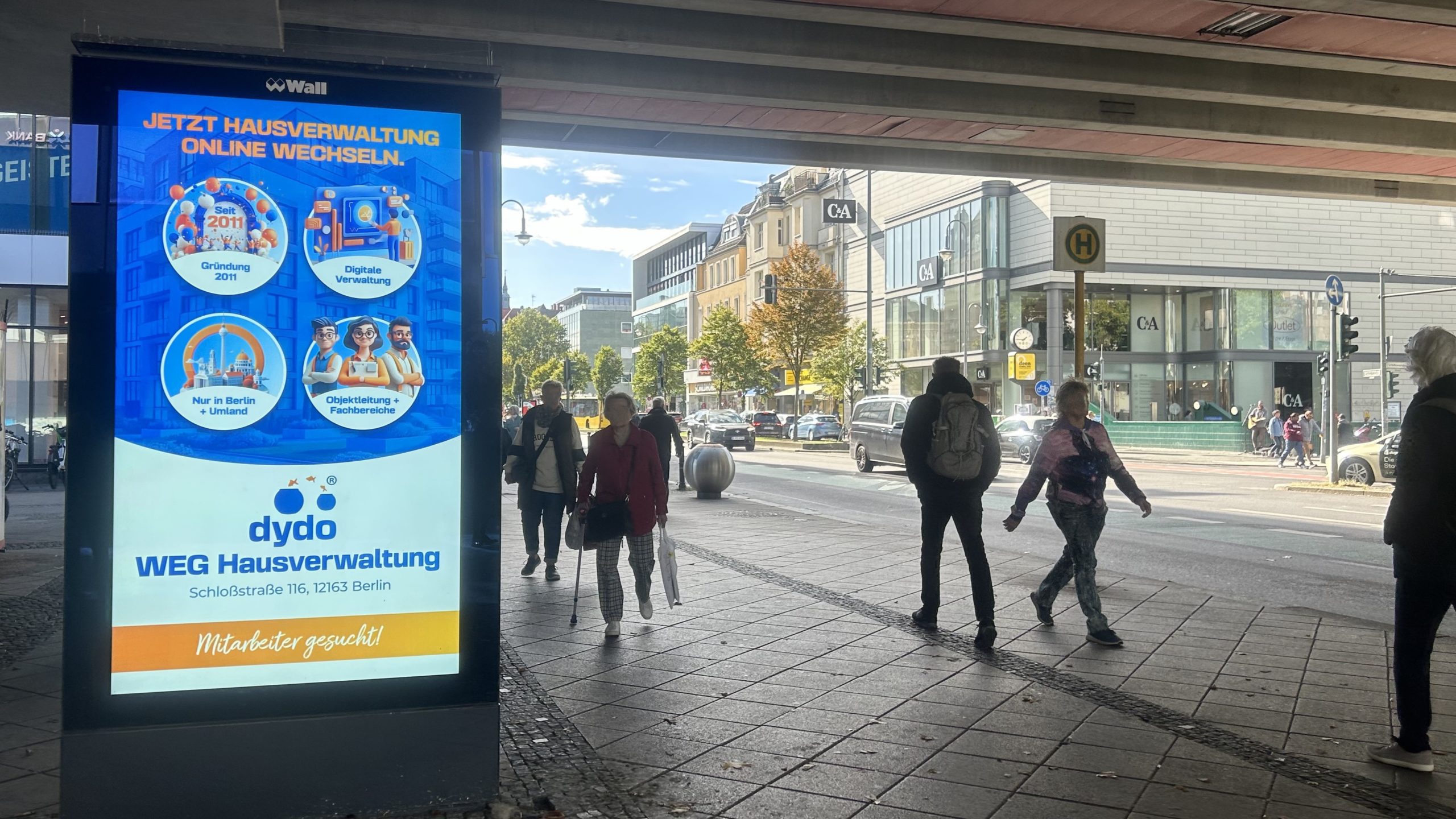 Auf der Schloßstraße in Berlin-Steglitz steht ein digitaler Screen auf dem Gehweg und strahlt gerade den Spot von Dydo aus. Die Immobilien & Hausverwaltung wirbt für neue Mitarbeiter. A 2 m² digital advertising medium is integrated into a roadside bus stop under a bridge in Berlin-Steglitz. The digital out-of-home advertising shows the property management company Dydo, which is looking for new employees.