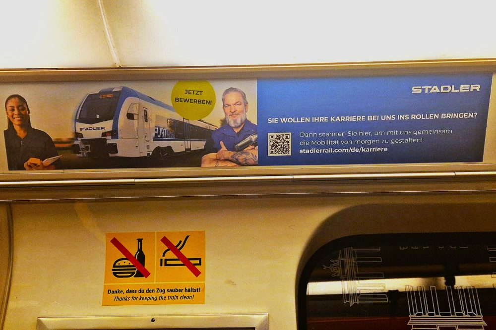 Auf der Nahaufnahme einer U-Bahn Innenwerbung sieht man einen Zug der Firma Stadler, sowie zwei Menschen. Der daneben platzierte Text erläutert, dass die Firma Stadler neue Mitarbeiter sucht. Side strip in the Berlin subway. It displays advertising from Stadler.