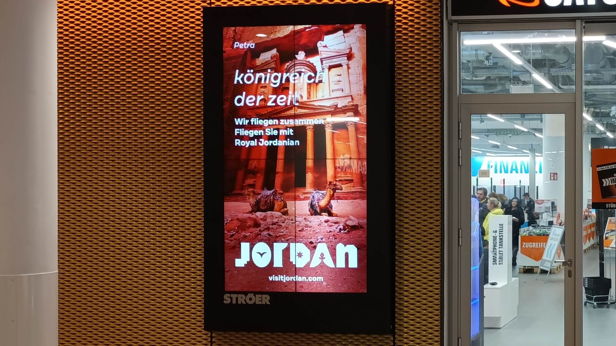 An der Wand in einer Mall hängt ein digitales City Light Poster, welches ein touristisches Motiv von Jordanien zeigt. Im rechten Teil des Bildes sieht man im Hintergrund ein Geschäft und Menschen.  