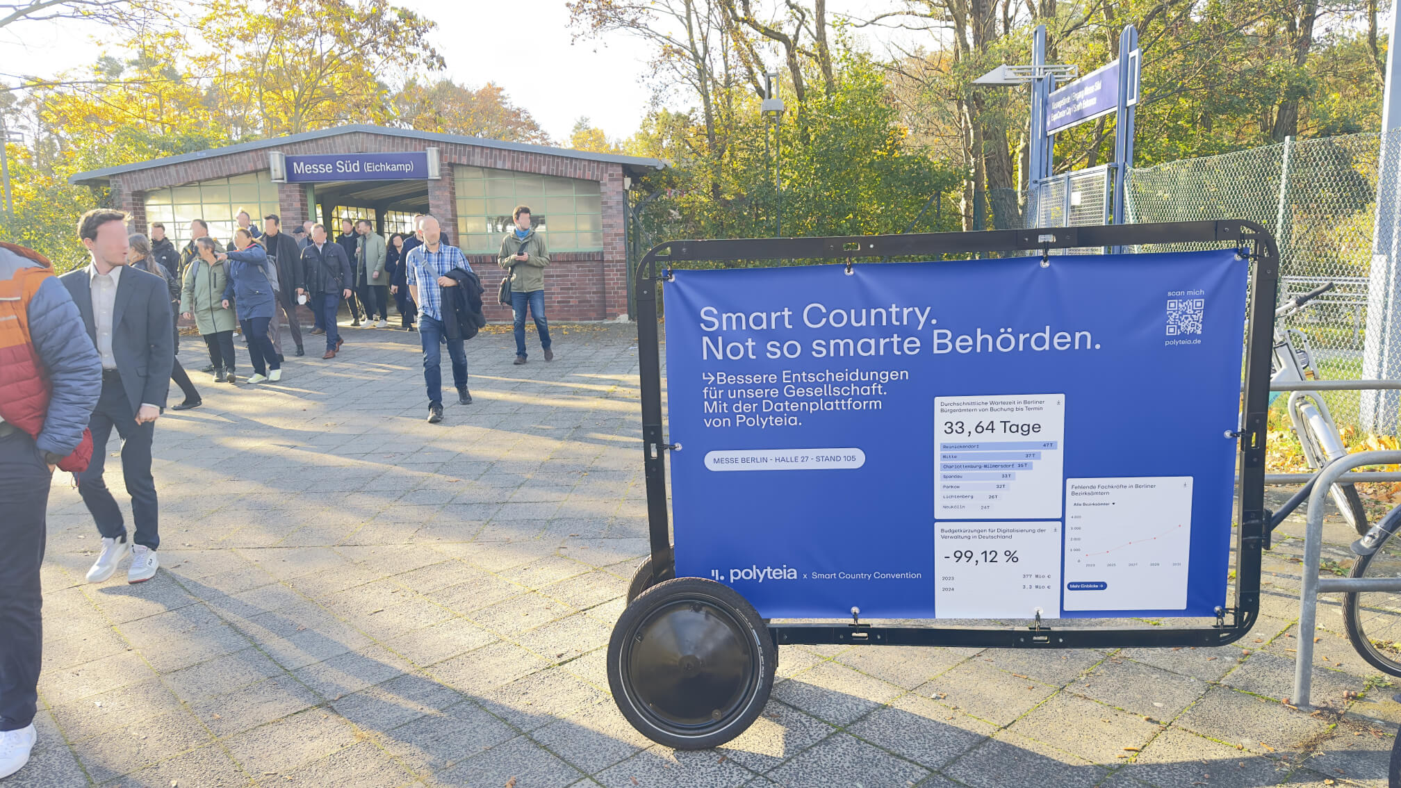 Vor dem Bahnhof Messe Süd steht ein Cool Lite Bike mit Werbeanhänger. Auf dem in einem Rahmen gespannten Banner sieht man die Werbung von Polyteia zur Smart Country Convention. Im Hintergrund strömen die Messebesucher aus dem Bahnhof in Richtung Messe.