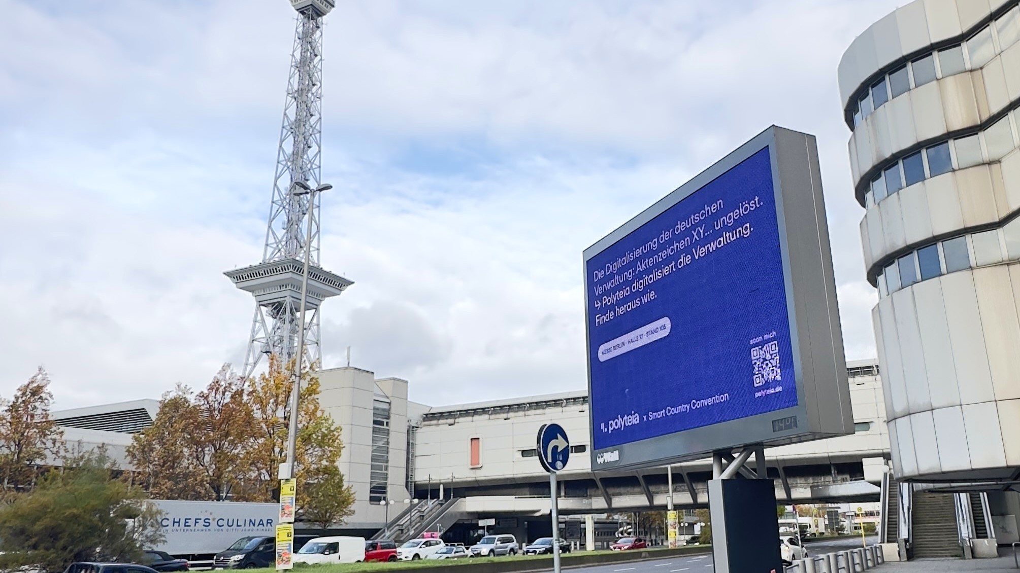 Am Messedamm in Berlin steht ein digitales City Light Board, welches die Werbung Polyteia zur Smart City Convention zeigt. Im Hintergrund sieht man die Messe Berlin und den Berliner Funkturm.