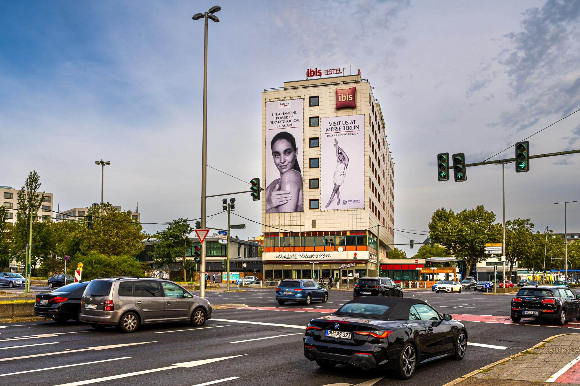 Am stark befahrenen Messedamm steht das IBIS Hotel. An der Fassade hängen zwei Riesenposter mit Werbung zur Messe.