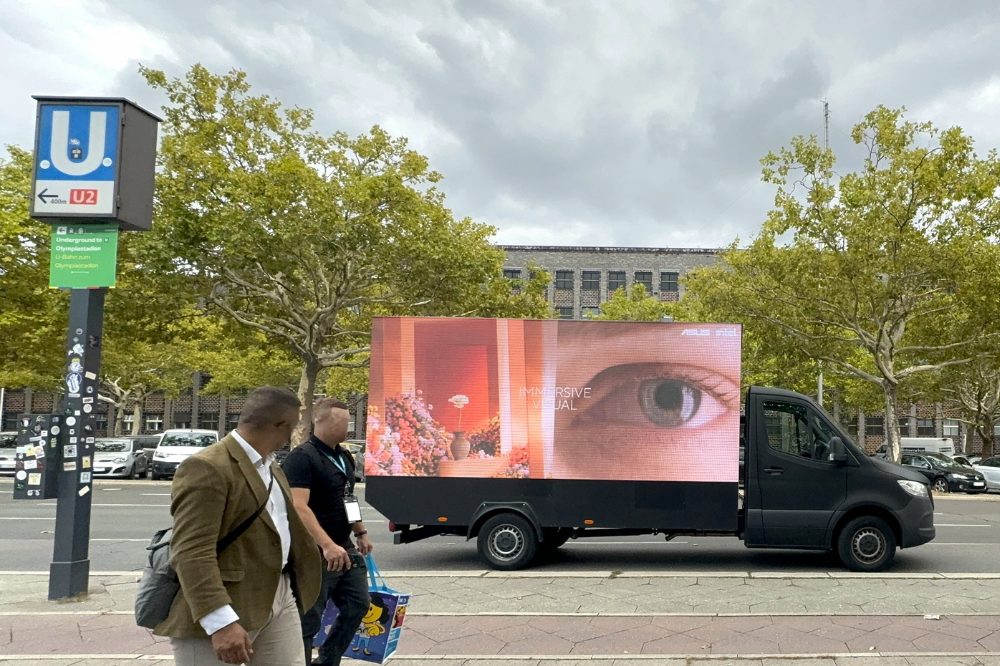 An der Straße vor dem Messegelände in Berlin, steht ein Video Truck, der auf seinen drei Screens einen Clip von ASUS zeigt. Davor laufen Menschen entlang und an der linken Seite ist ein Schild der U-Bahn (U2). Hinter dem digitalen Werbeträger stehen Bäume, Autos und Gebäude. In front of the IFA trade fair grounds in Berlin, the video truck showing an advertisement from ASUS is driving around. Walking past the truck are two people. In the background there are cars, threes and buildings.