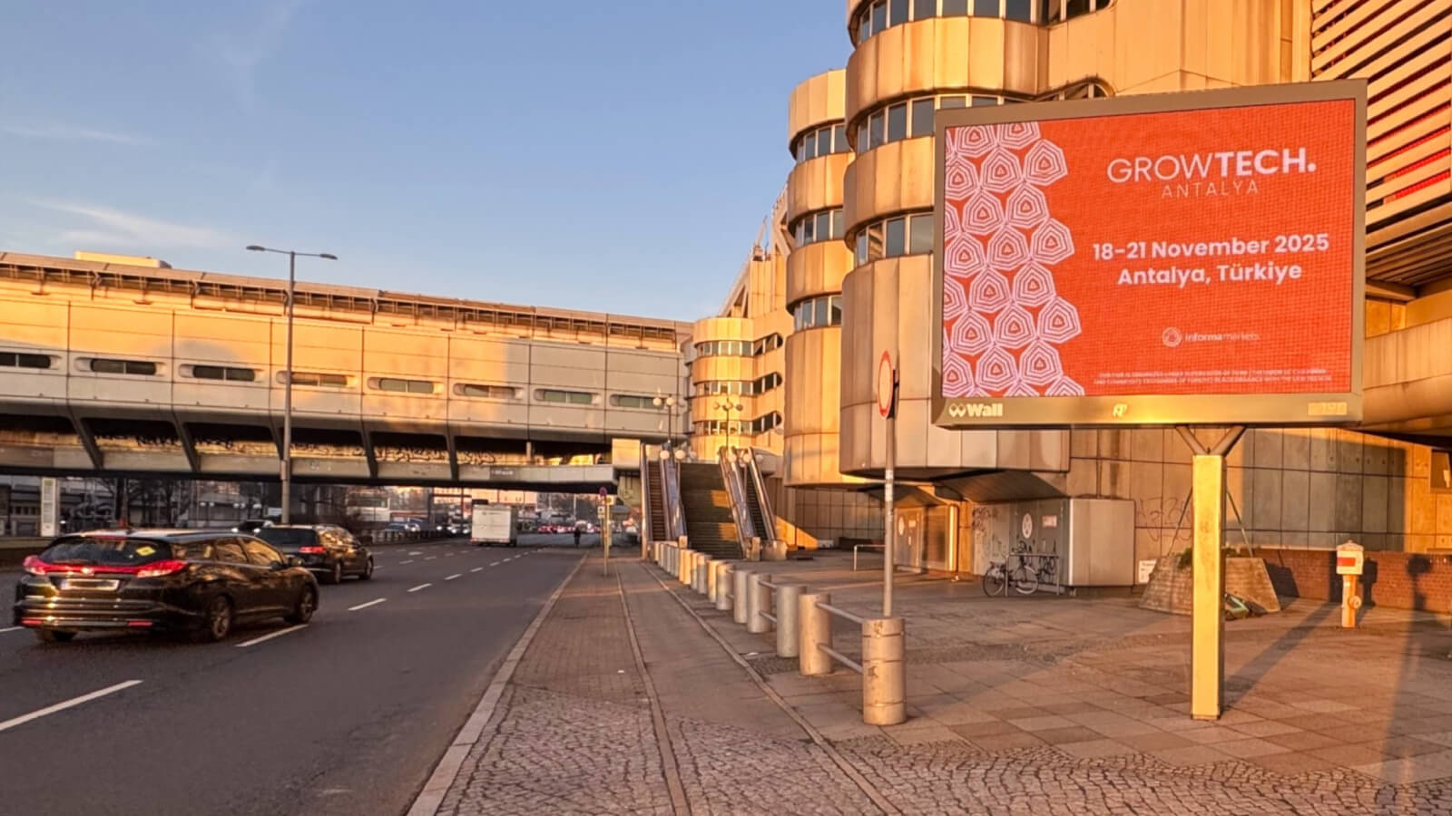 Am stark befahrenen Messedamm in Berlin steht ein digitales City Light Board. Auf dem digitalen Bildschirm sieht man in knalligem Rot eine Eventankündigung. Im Hintergrund sieht man Teile der Messegeländes und das ICC.