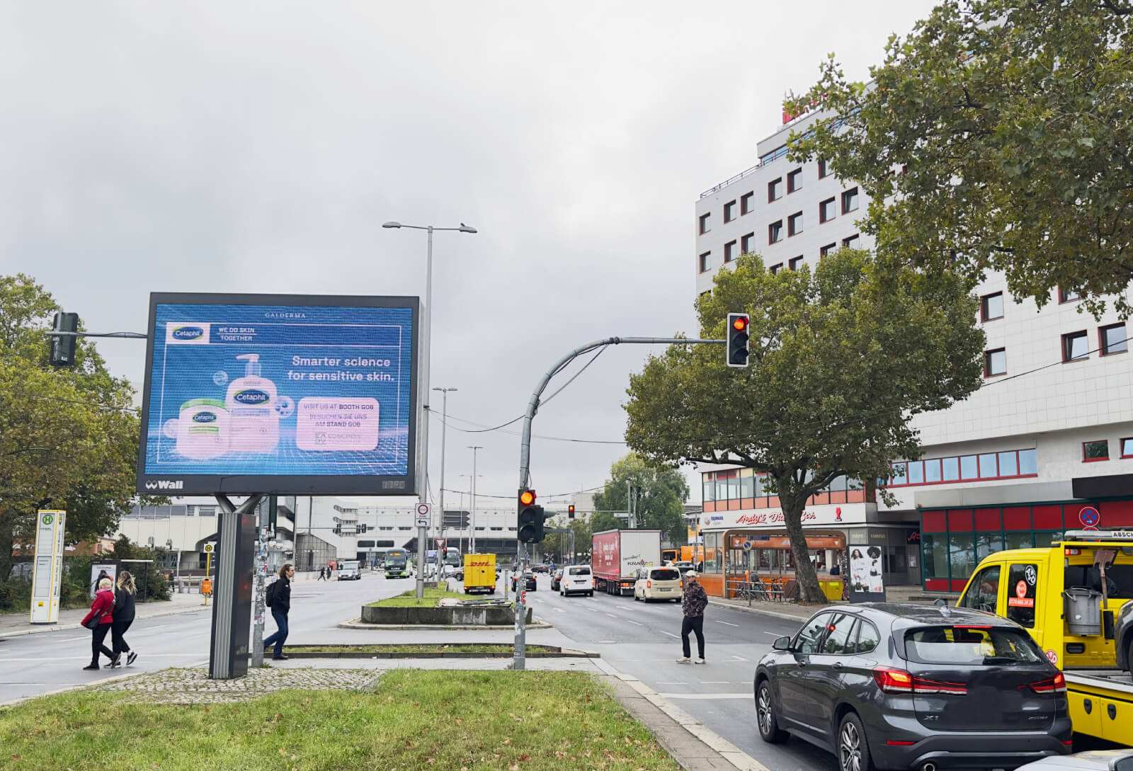 An einer Ampel in Berlin stehen wartende Fahrzeuge und Fußgänger überqueren die Straße. Auf dem Grünstreifen in der Mitte steht ein digitales City Light Board auf dem die Werbung der Kosmetikfirma Galderma zu sehen ist.